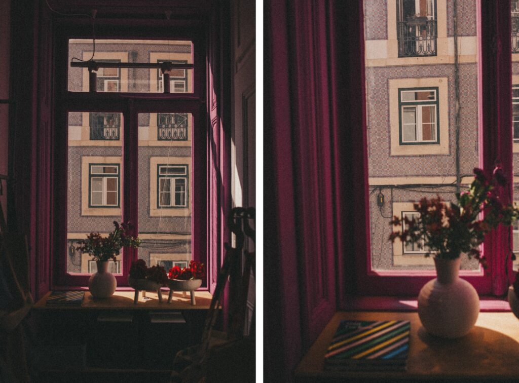 Ein sonnenbeschienenes Fenster mit magentafarbenen Rahmen blickt auf ein gemustertes Gebäude. Auf der Fensterbank stehen Blumen in einer weißen Vase neben bunten Büchern - eine einladende Szene, die ein Familien Fotograf in einem Fotostudio Köln oft einfängt.