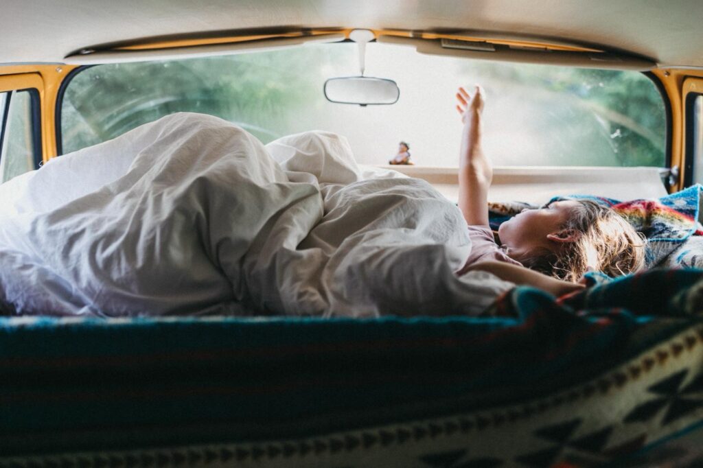 Eine Person liegt unter einer weißen Decke auf dem Rücksitz eines gelben Vans und streckt den Arm in die Morgensonne. Wie ein Kinderfotograf in Köln diesen entspannten, gemütlichen Moment einfängt, füllt das weiche natürliche Licht die Szene mit Wärme.