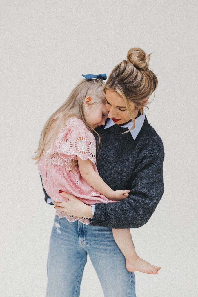 Eine Frau in einem dunklen Pullover und Jeans hält ein junges Mädchen in einem rosa Spitzenkleid mit blauer Schleife, beide lächeln und umarmen sich vor einem schlichten weißen Hintergrund - aufgenommen von einem erfahrenen Familienfotografen.