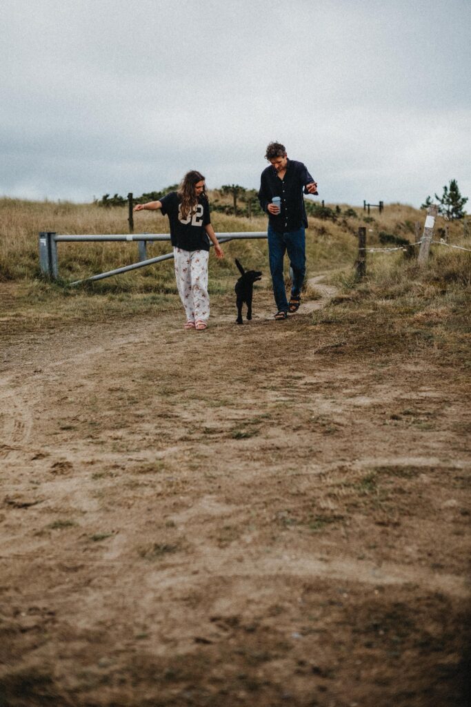 Ein Mann und ein junges Mädchen gehen mit einem kleinen schwarzen Hund einen Feldweg entlang. Aufgenommen von einem talentierten Kinderfotografen, genießen die beiden die freie Natur in einer grasbewachsenen ländlichen Gegend unter einem bewölkten Himmel, wobei das Mädchen einen Pyjama und ein Sporttrikot trägt.