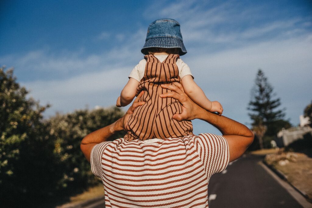 Eine Person in einem gestreiften Hemd trägt ein kleines Kind auf den Schultern. Das von einem Werbefotografen aufgenommene Kind mit einem blauen Eimerhut genießt den sonnigen Tag im Freien, während sie eine von Grünanlagen gesäumte Straße entlanggehen.