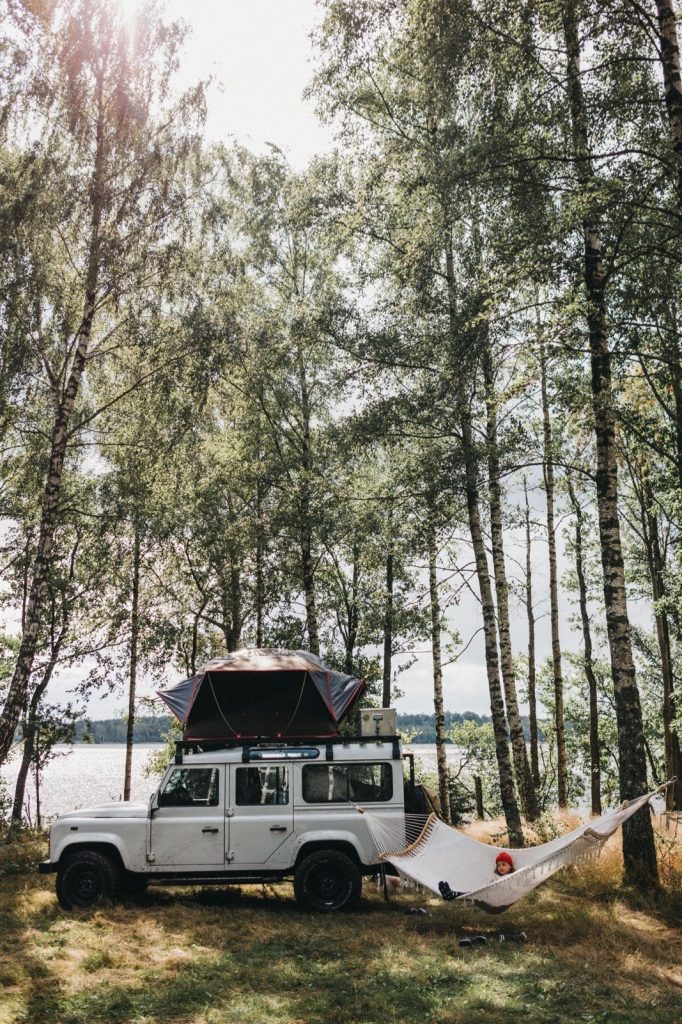 Ein weißer Geländewagen mit einem Dachzelt und einer Hängematte parkt zwischen hohen Bäumen in der Nähe eines Sees, das Sonnenlicht bricht durch die Blätter, perfekt eingefangen von einem Familienfotografen für eine unvergessliche Campingszene.