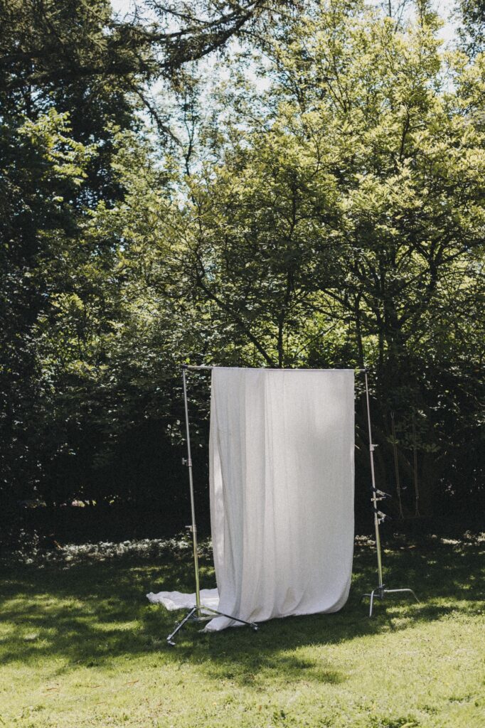 Ein weißes Laken hängt an einem Metallrahmen mitten auf einer Wiese, umgeben von hohen grünen Bäumen und schummrigem Sonnenlicht - eine einladende Szene für jeden Fotografen Köln und jede kreative Fotografin auf der Suche nach einzigartiger Inspiration.