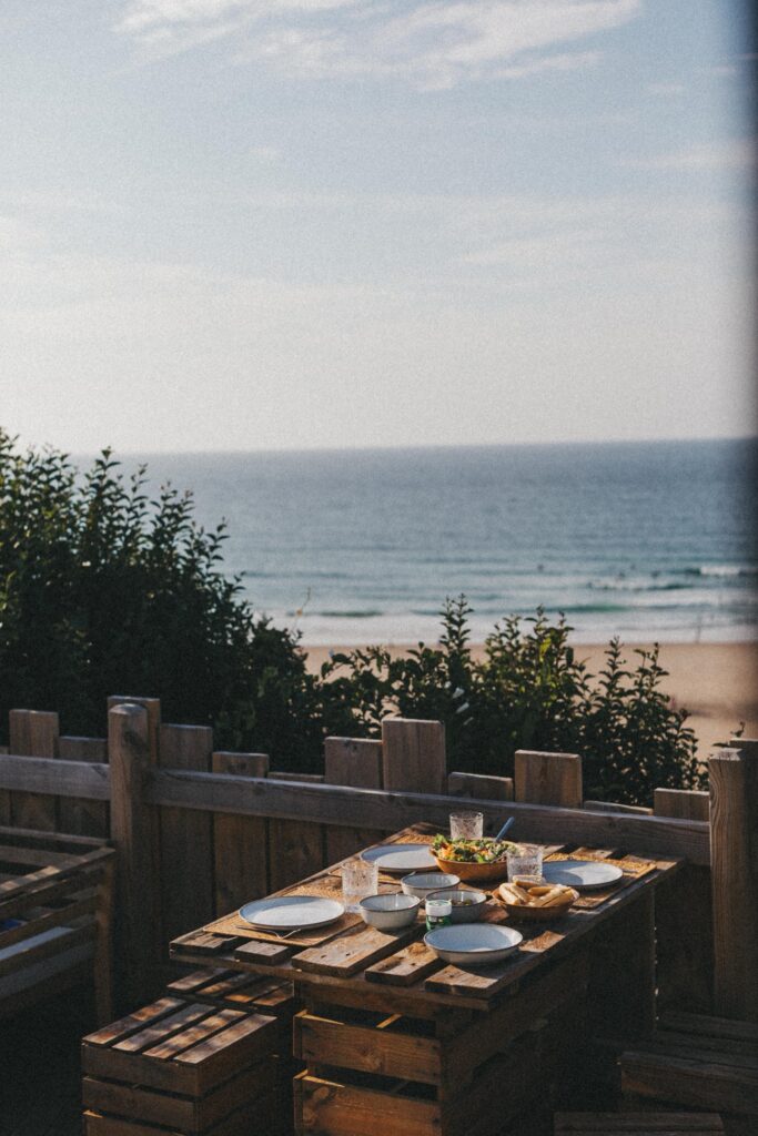 Ein rustikaler Holztisch mit Tellern, Gläsern und Speisen steht auf einer Terrasse mit Blick auf das Meer. Das Sonnenlicht beleuchtet die Szene - und fängt den perfekten Moment ein, wie durch die Linse eines talentierten Fotografen Kölns gesehen.