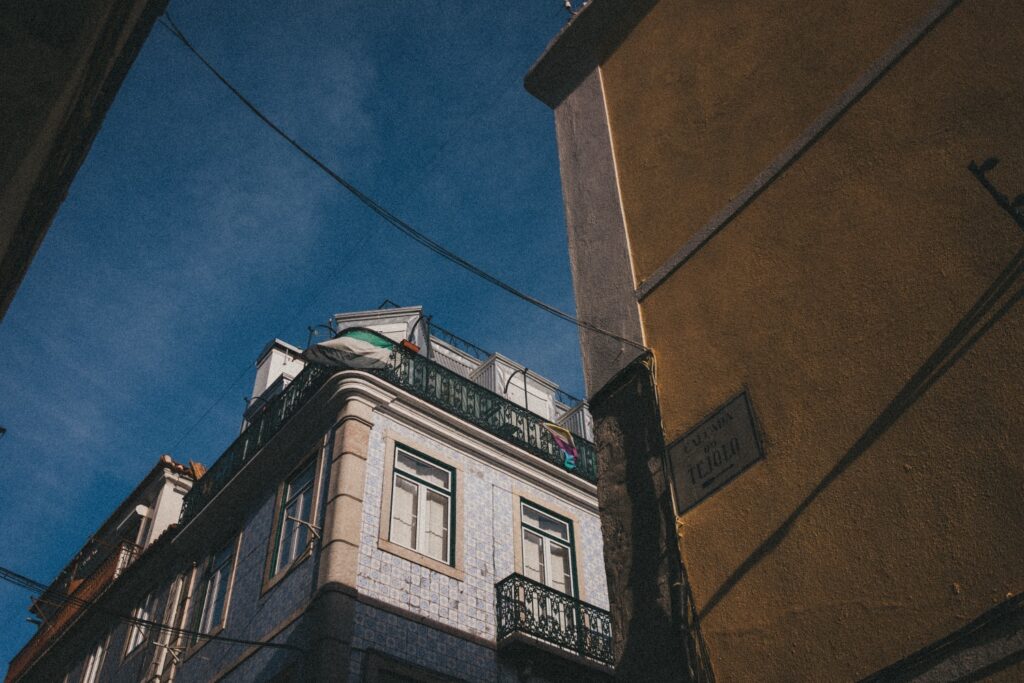 Blick aus geringer Höhe auf ein Eckhaus mit blau gemusterten Fliesen und grünen Balkonen vor einem klaren Himmel. Gelbe und graue Wände rahmen die Szene ein, die von einem Werbefotografen aufgenommen wurde, während sich Stromleitungen über dem Gebäude kreuzen.