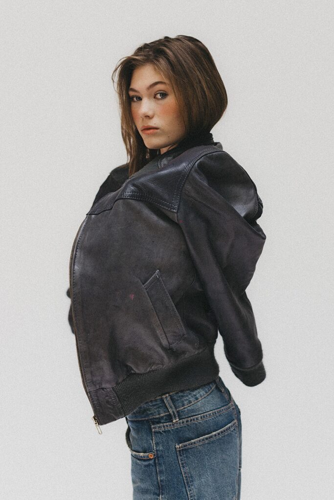 Eine junge Frau mit braunen Haaren, gestylt wie ein Familien Fotograf, trägt eine lockere, dunkle Lederjacke und blaue Jeans, steht seitlich vor einem schlichten weißen Hintergrund und schaut in die Kamera.