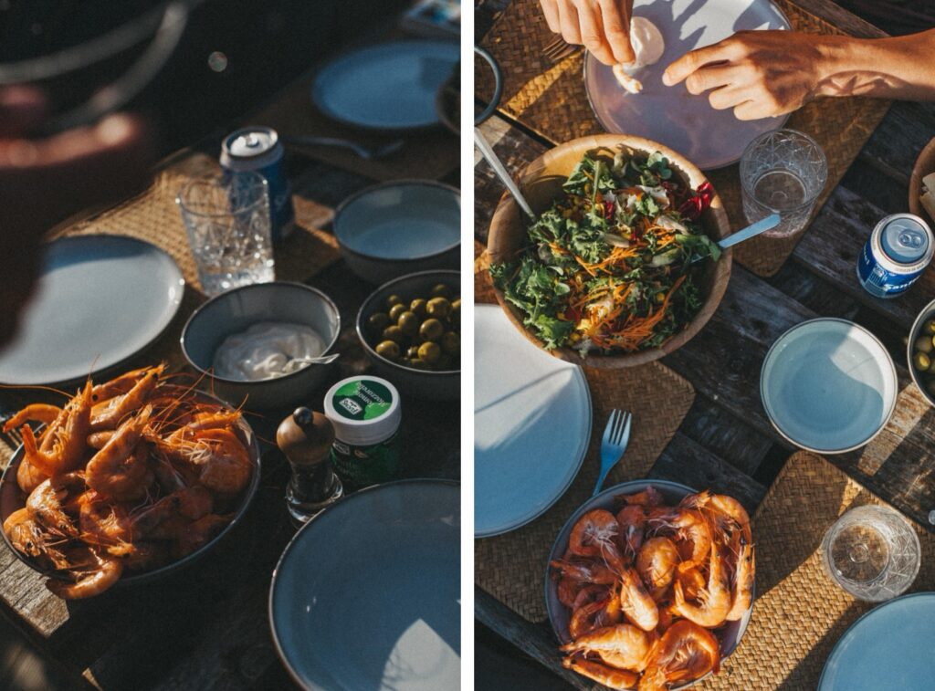 Ein im Freien gedeckter Tisch mit Tellern, Schalen mit Garnelen, Salat, Oliven und Getränken. Die Hände greifen nach dem Essen, während das Sonnenlicht Schatten auf den rustikalen Holztisch wirft - eingefangen von einem talentierten Werbefotografen für ein authentisches Esserlebnis.