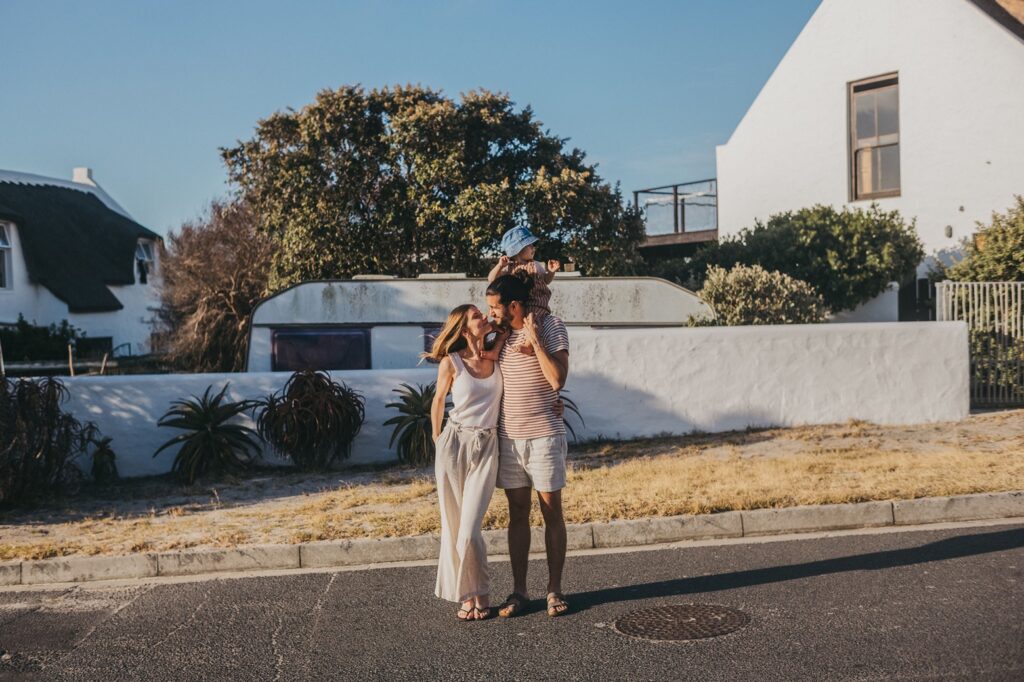 Ein lächelndes Paar steht zusammen auf einer sonnigen Straße, wobei der Mann ein Kind auf seinen Schultern trägt. Aufgenommen von einem talentierten Kinderfotografen, genießen sie einen ungezwungenen, entspannten Moment im Freien vor weißen Häusern und Grünanlagen.