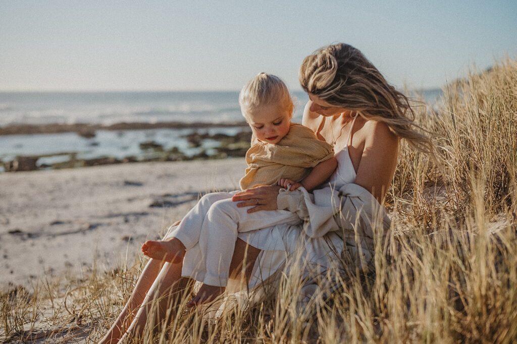 Eine Frau sitzt im hohen Strandgras und hält ein kleines Kind, das in eine gelbe Decke eingewickelt ist. Aufgenommen von einer erfahrenen Fotografin, blicken sie liebevoll nach unten, mit dem Meer und den Felsen im Hintergrund unter einem klaren Himmel.