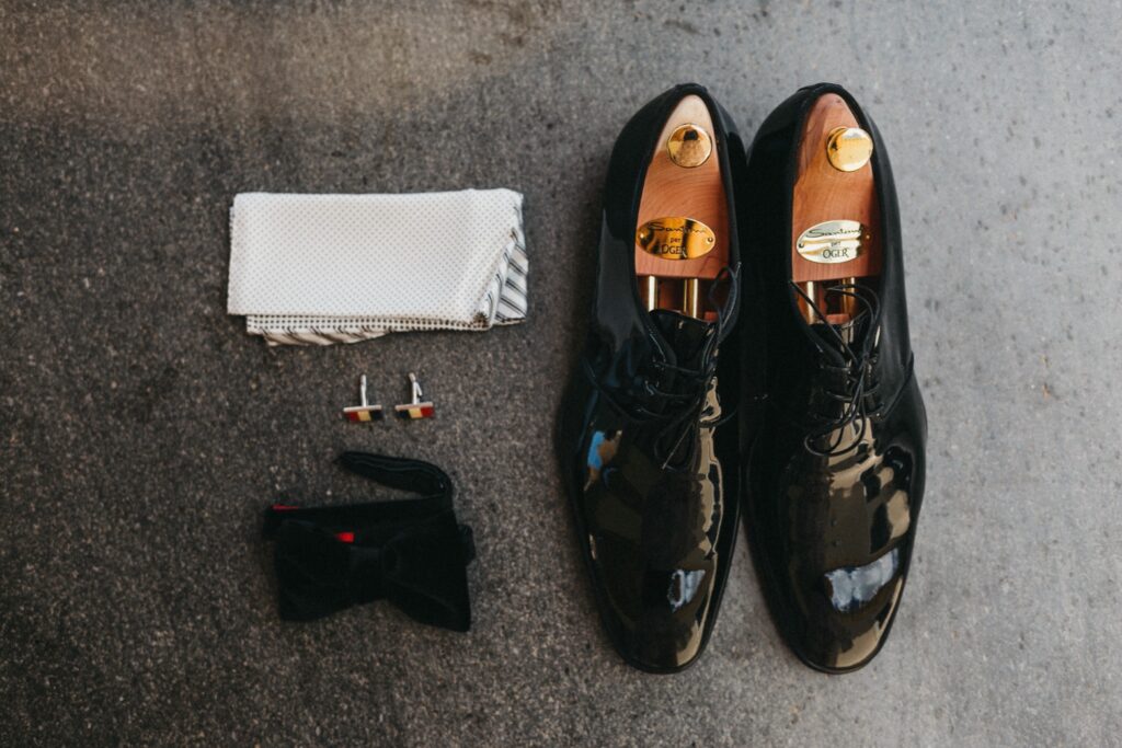 Glänzende schwarze Anzugschuhe, ein gefaltetes weißes Einstecktuch, Manschettenknöpfe und eine schwarze Fliege, ordentlich auf einer grauen, strukturierten Oberfläche angeordnet - perfekt für eine elegante sizilianische Hochzeit.