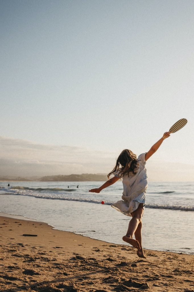 Eine Frau in einem weißen Kleid spielt Paddelball an einem Sandstrand in der Nähe der Küste bei Sonnenuntergang - Wellen, entfernte Schwimmer und ein dunstiger Himmel bilden die Kulisse, perfekt für jeden Fotograf Köln oder Kinderfotograf, der natürliche, ehrliche Momente sucht.