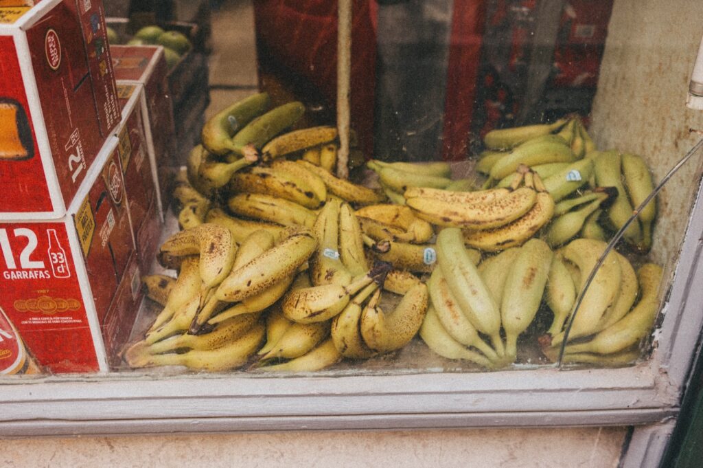 Ein Stapel reifer und leicht überreifer Bananen liegt hinter einer Glasscheibe, daneben stapeln sich rote Kartons mit der weißen Aufschrift "12" - perfekt für einen Werbefotografen oder ein Fotostudio Köln, das lebendige, authentische Bilder sucht.