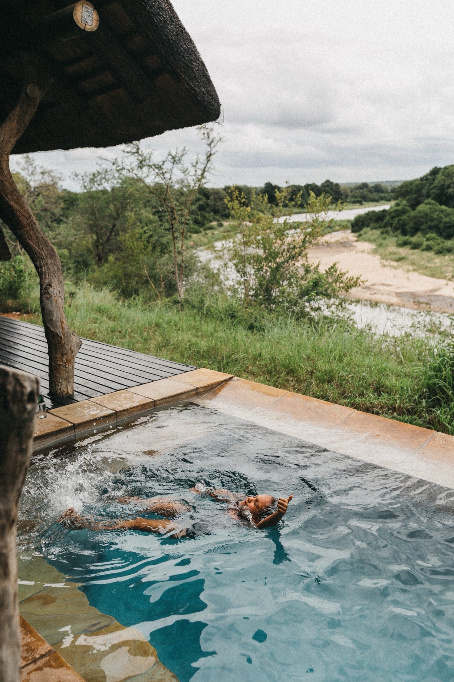 Eine Person schwimmt in einem kleinen Außenpool an einer rustikalen Holzkonstruktion, umgeben von Grün und mit Blick auf einen Fluss - eingefangen von einem Werbefotografen, wobei Bäume und Wolken die Stimmung bestimmen.