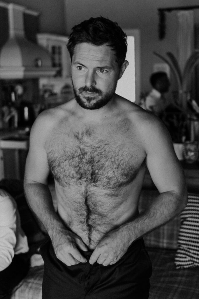 Ein bärtiger Mann ohne Hemd steht in einem Haus und schaut zur Seite, während er seine Hose zurechtrückt. Das Schwarz-Weiß-Bild zeigt eine sizilianische Hochzeitsszene in einer Küche und einem Wohnbereich, wobei andere Personen teilweise im Hintergrund zu sehen sind.