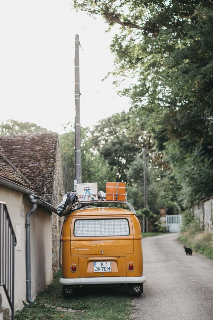 Ein gelber Oldtimer-Kastenwagen mit Gepäck auf dem Dach ist auf einer Landstraße neben einem rustikalen Haus geparkt, das von Bäumen umgeben ist. Eine schwarze Katze läuft voraus. Diese ruhige Szene, die von einem Familienfotografen aufgenommen wurde, könnte aus einem Portfolio des Fotostudios Köln stammen.