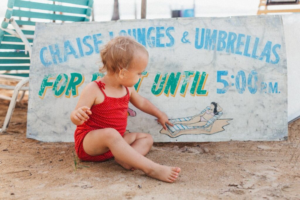 Ein Kleinkind in einem roten Outfit sitzt im Sand und zeigt auf ein verwittertes Schild für Liegestühle und Sonnenschirme. Aufgenommen von einer talentierten Fotografin aus dem Fotostudio Köln, vervollständigen grüne und weiße Stühle die entspannte Strandszene.