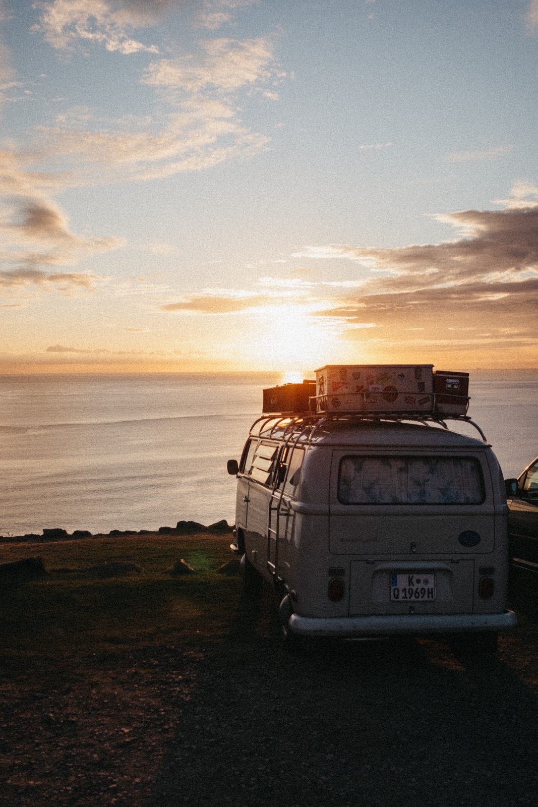 Ein Oldtimer-Van mit Gepäck auf dem Dach parkt bei Sonnenuntergang am Meer, mit Blick auf ruhiges Wasser und einen leuchtenden Himmel. Diese Szene, die von einem talentierten Fotografen aus Köln aufgenommen wurde, weckt Nostalgie und Abenteuerlust.