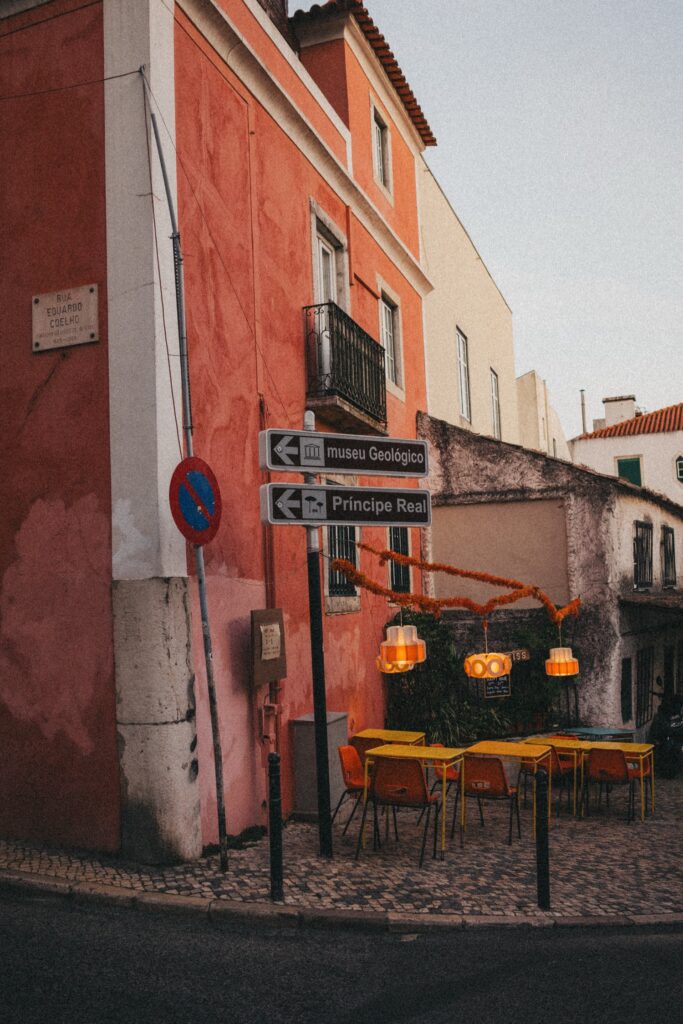 Ein gemütliches Straßencafé mit gelben Tischen und Stühlen liegt an einer kopfsteingepflasterten Straßenecke, direkt neben einem rot-weißen Gebäude. Über dem Sitzbereich hängen orangefarbene Lichter - perfekt für einen Werbefotografen oder Kinderfotografen, der lebendige, urbane Szenen sucht.