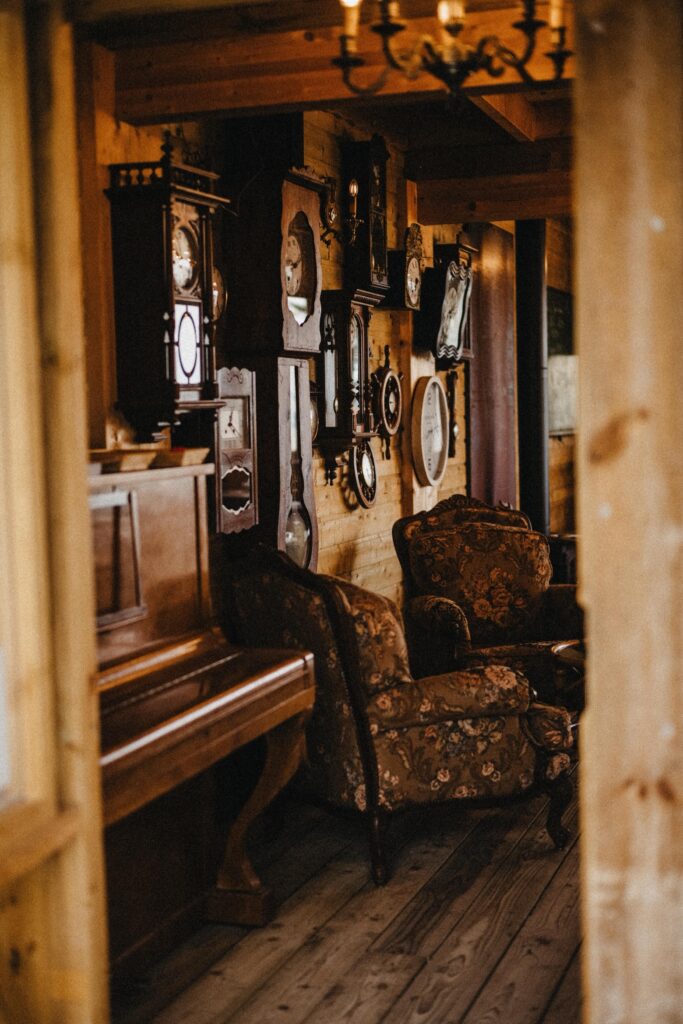 Ein gemütlicher Vintage-Raum mit einem geblümten Sessel, einem Klavier und einem Holzboden. Die Wände sind mit antiken Uhren und Spiegeln geschmückt und bieten eine ideale Kulisse für einen Kinder- oder Familienfotografen, der eine warme, nostalgische Atmosphäre sucht.