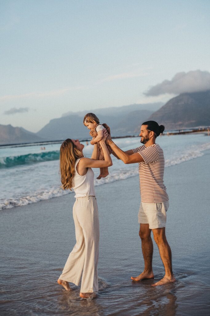 Ein lächelndes Paar steht barfuß an einem Strand und hebt spielerisch sein kleines Kind zwischen sich. Aufgenommen von einem Familienfotografen, erscheinen Wellen und ferne Berge im Hintergrund unter einem teilweise bewölkten Himmel.