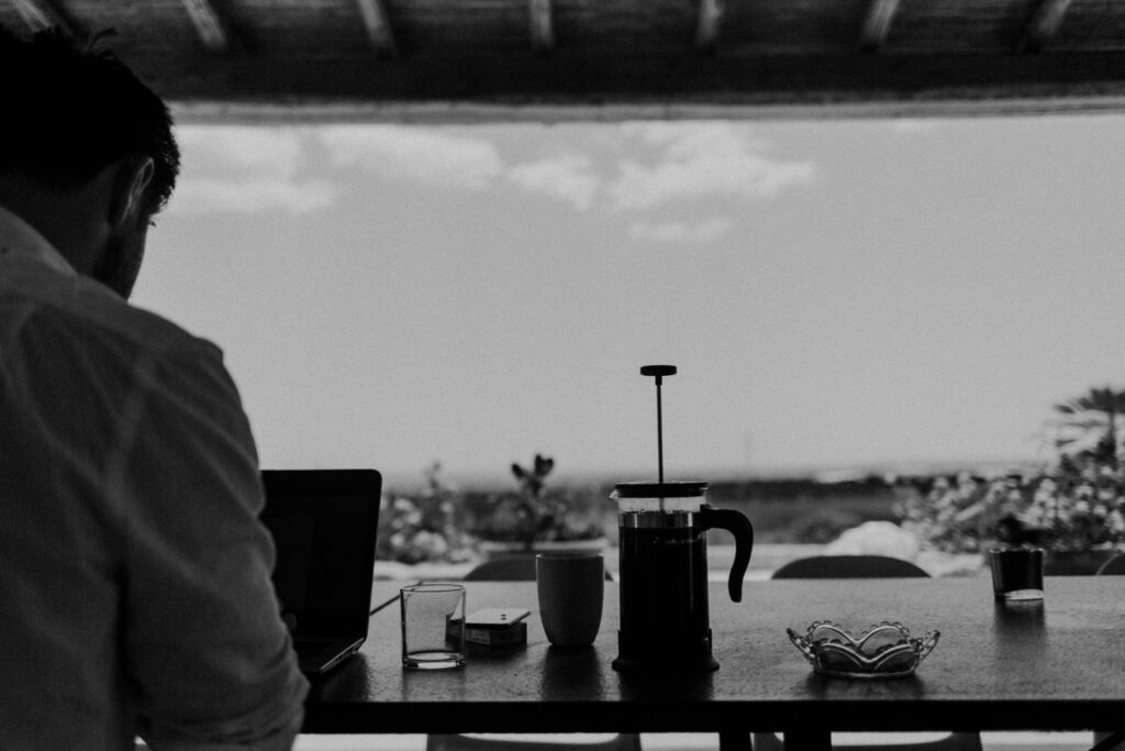 Eine Person sitzt an einem Tisch und arbeitet an einem Laptop, daneben stehen eine Kaffeemaschine, eine Tasse, ein Glas und ein Aschenbecher. Im Hintergrund ist der Blick nach draußen und der helle Himmel zu sehen - ein ruhiger Moment vor einer Hochzeit in Sizilien. Das Foto ist in schwarz-weiß gehalten.