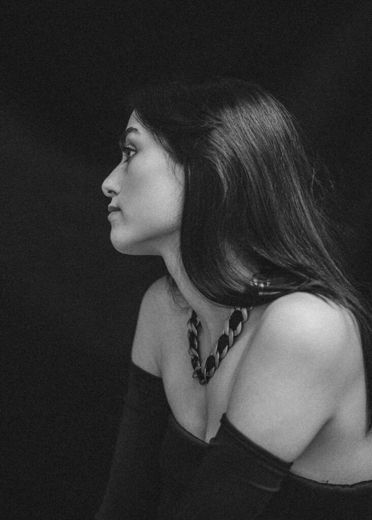 Schwarz-Weiß-Foto von Fotografin Köln einer Frau mit langen dunklen Haaren, die ein schulterfreies Oberteil und eine klobige Halskette trägt und im Profil vor einem dunklen Hintergrund posiert.