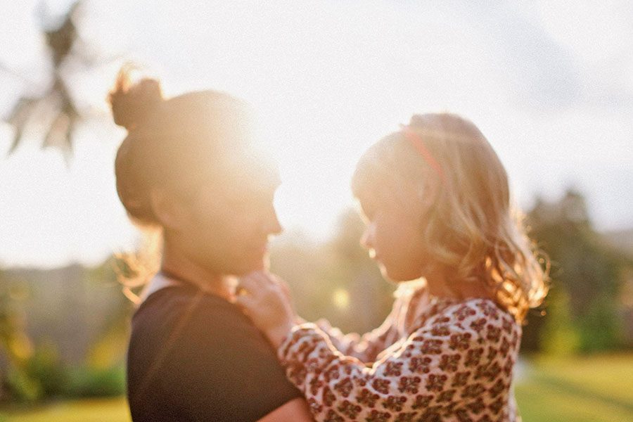 Ein verschwommenes, sonnenbeschienenes Foto eines Familienfotografen zeigt einen Erwachsenen und ein kleines Kind, die sich im Freien gegenüberstehen. Das Kind hält sanft das Hemd des Erwachsenen, während das Sonnenlicht hinter ihnen leuchtet und eine warme und strahlende Atmosphäre schafft.