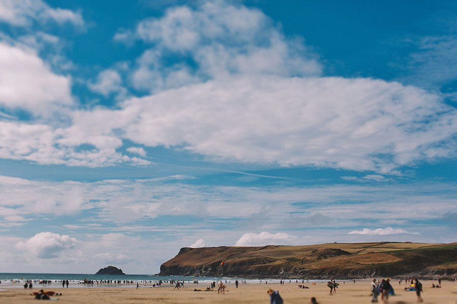 Ein Sandstrand mit verstreuten Menschen, die sich entspannen und spazieren gehen, unter einem strahlend blauen Himmel mit großen weißen Wolken. In der Ferne erheben sich grasbewachsene Hügel und Klippen - eine ideale Kulisse für eine Kids Fotograf oder Fotostudio Köln Session.