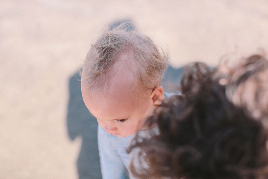 Nahaufnahme eines jungen Kindes mit feinem, hellblondem Haar, das von oben nach unten schaut. Ein Teil einer anderen Person mit lockigen braunen Haaren ist im weich verschwommenen Hintergrund zu sehen. Aufgenommen von einem Fotograf Köln in einem professionellen Fotostudio Köln.