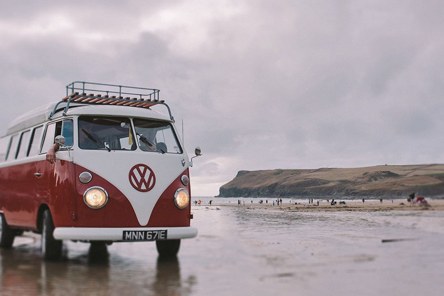 Ein rot-weißer Oldtimer-Volkswagen parkt an einem nassen Strand, aufgenommen von einem Werbefotografen, mit Menschen in der Ferne und einer grasbewachsenen Klippe unter einem bewölkten Himmel im Hintergrund.
