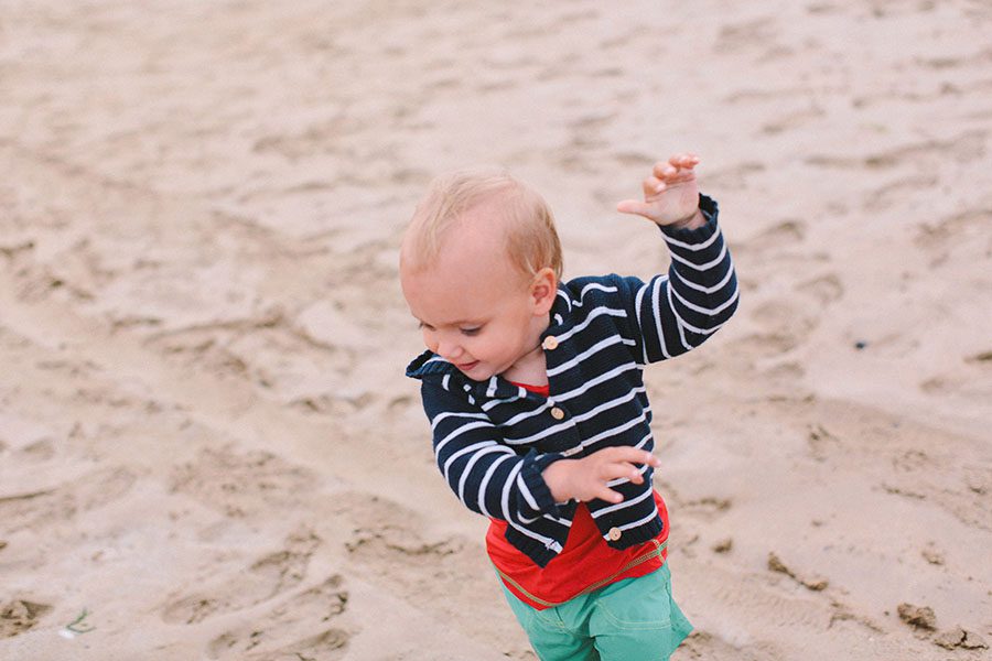 Ein kleines Kind, das von einem talentierten Kinderfotografen aufgenommen wurde, trägt eine gestreifte Jacke, ein rotes Hemd und eine grüne Hose, während es an einem Sandstrand spielt, einen Arm hebt und mit einem freudigen Ausdruck nach unten schaut.