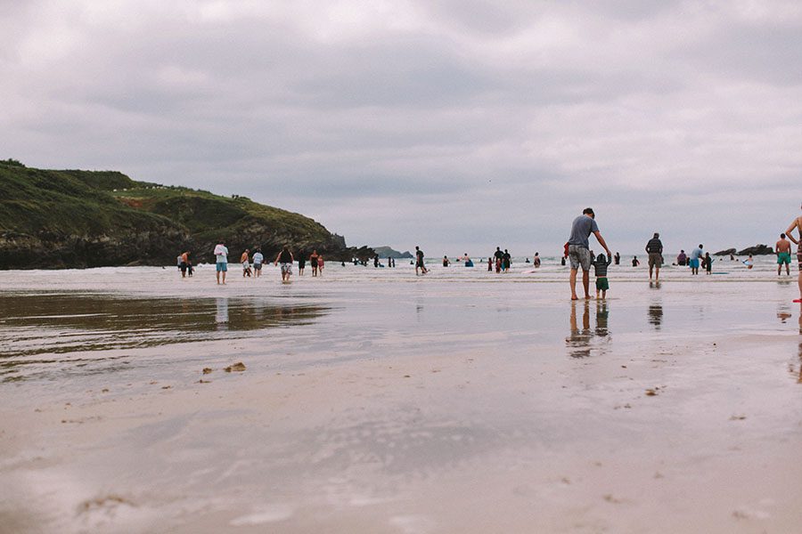 Menschen spazieren und waten an einem Sandstrand unter einem bewölkten Himmel, wobei sich ihre Figuren im flachen Wasser spiegeln. Im Hintergrund sind grüne Klippen zu sehen. Diese ruhige Szene inspiriert jeden Werbefotografen, der natürliche, entspannte Momente sucht.