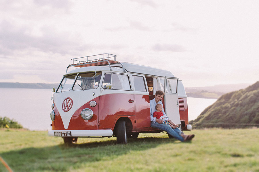 Ein klassisches rot-weißes Volkswagen Wohnmobil steht auf einem Grashügel mit Blick auf das Wasser. Aufgenommen von einem talentierten Kinderfotografen, sitzen ein Erwachsener und ein Kind in der offenen Seitentür und genießen die Aussicht bei bewölktem Himmel.