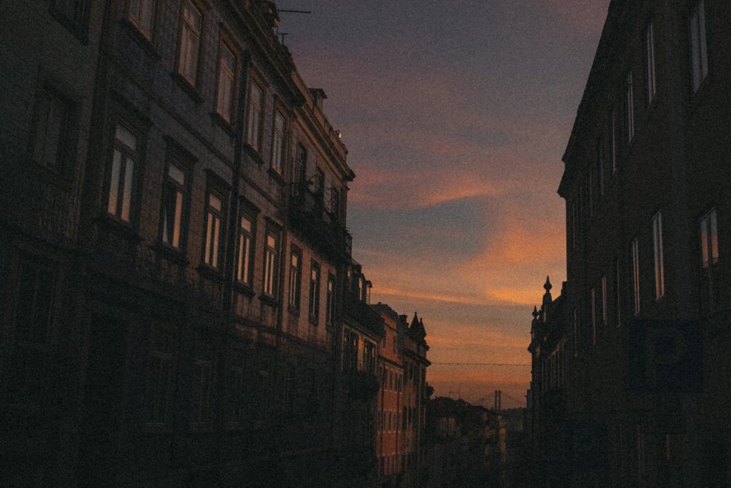 Hohe Gebäude bilden eine enge Gasse bei Sonnenuntergang, rosa und orangefarbene Wolken erhellen den Himmel, während die Fenster das schwindende Licht reflektieren - die perfekte Inspiration für einen Fotografen Köln, der Stadtszenen oder Momente als Familienfotograf festhält.