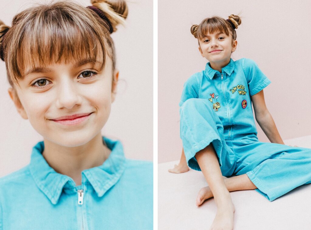 Ein junges Mädchen mit braunem Haar in einem doppelten Dutt, das einen hellblauen Overall mit bunten Aufnähern trägt, lächelt auf zwei Fotos eines Kinderfotografen in die Kamera - eine Nahaufnahme und eine sitzende Ganzkörperpose - vor einem hellrosa Hintergrund.