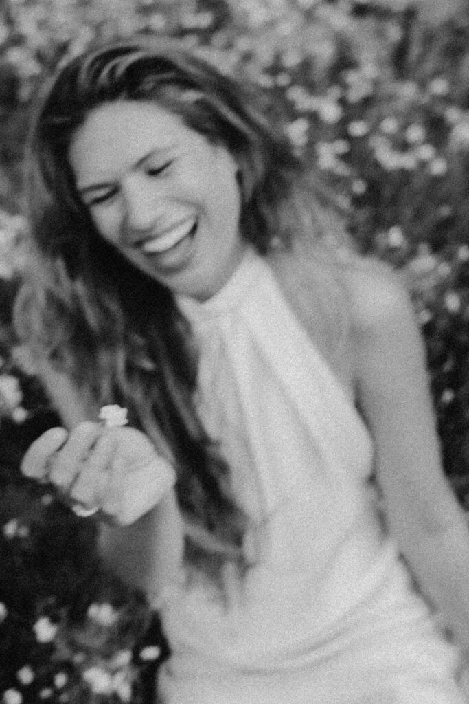 Schwarz-Weiß-Foto einer Frau mit langen Haaren und ärmellosem Kleid, die lächelnd und lachend eine kleine Blume hält. Die fröhliche Szene wurde von einem Werbefotografen aufgenommen und ist von unscharfem Laub und Blumen umgeben.