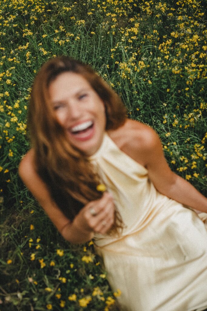Eine Frau in einem cremefarbenen Kleid sitzt in einem gelben Blumenfeld und lacht mit zurückgelegtem Kopf. Das Bild der Fotografin Köln ist leicht unscharf und vermittelt eine fröhliche und unbeschwerte Atmosphäre.