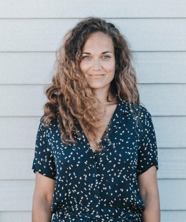 Eine Frau mit lockigen braunen Haaren und blauen Augen steht vor einer hellen Holzwand und lächelt leicht. Sie trägt eine marineblau gemusterte Bluse - ein authentischer Moment, der von einem Familienfotografen während eines Familienfotoshootings eingefangen wurde.