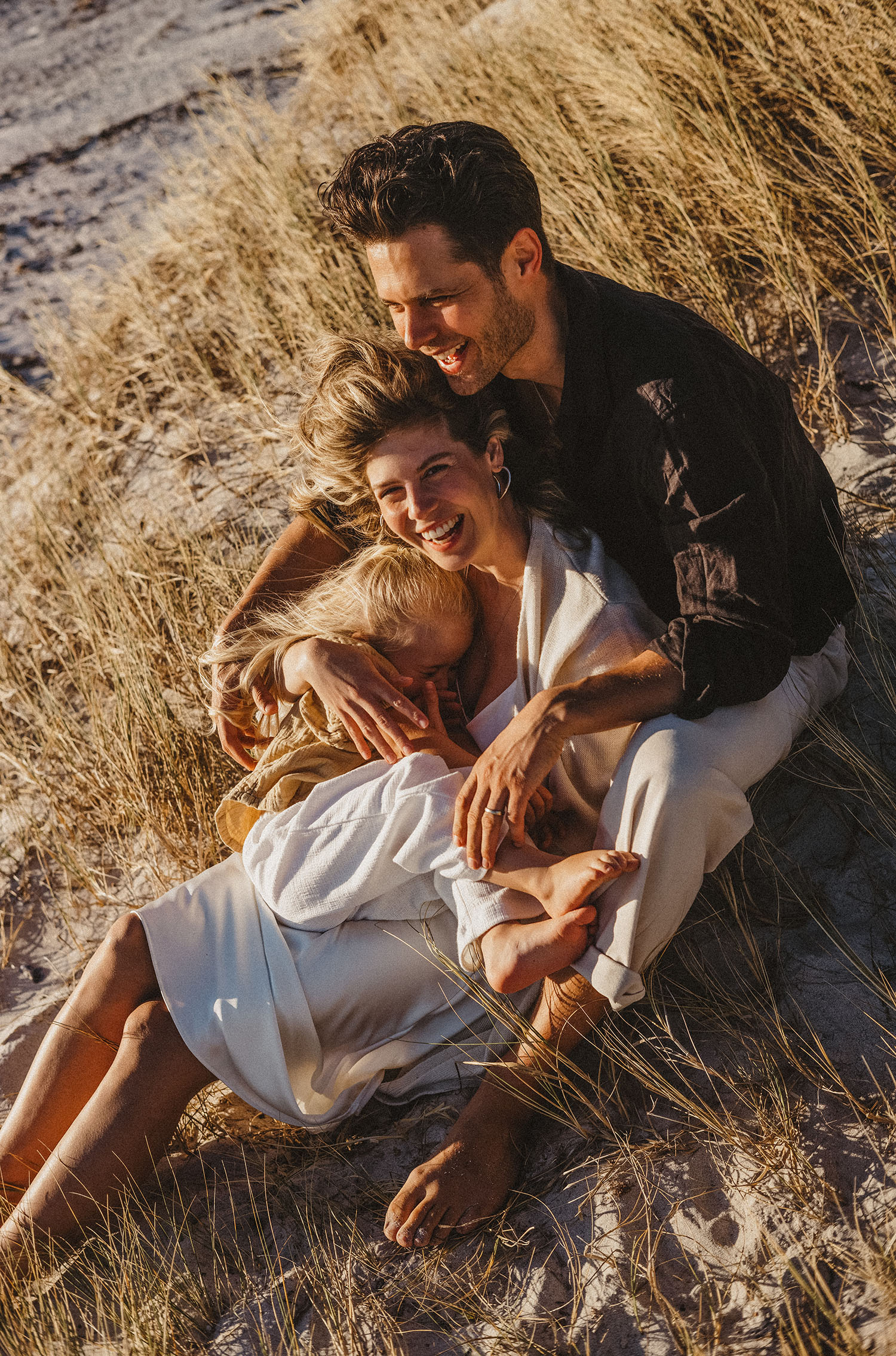 Ein glückliches Paar sitzt im hohen Strandgras, lächelt und umarmt ein kleines Kind, das in eine leichte Decke eingewickelt ist, während das warme Sonnenlicht - das von einer erfahrenen Fotografin gekonnt eingefangen wurde - einen goldenen Schein auf sie wirft.