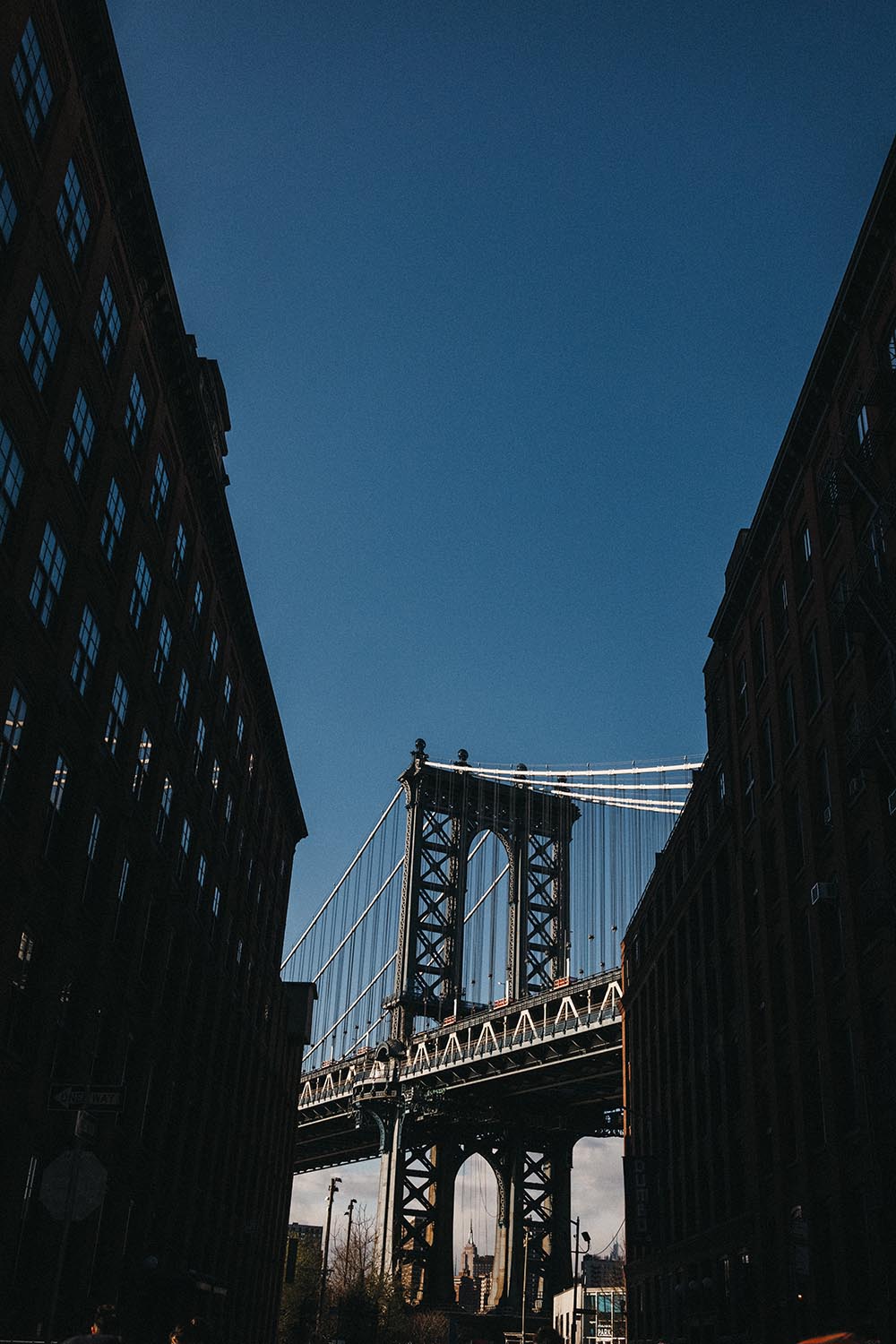 Die Manhattan Bridge liegt zwischen zwei hohen, schattigen Backsteingebäuden unter einem klaren blauen Himmel. Das Sonnenlicht hebt die Stahlkonstruktion in der Mitte hervor und inspiriert jeden Familienfotografen, der eine eindrucksvolle Stadtkulisse sucht.