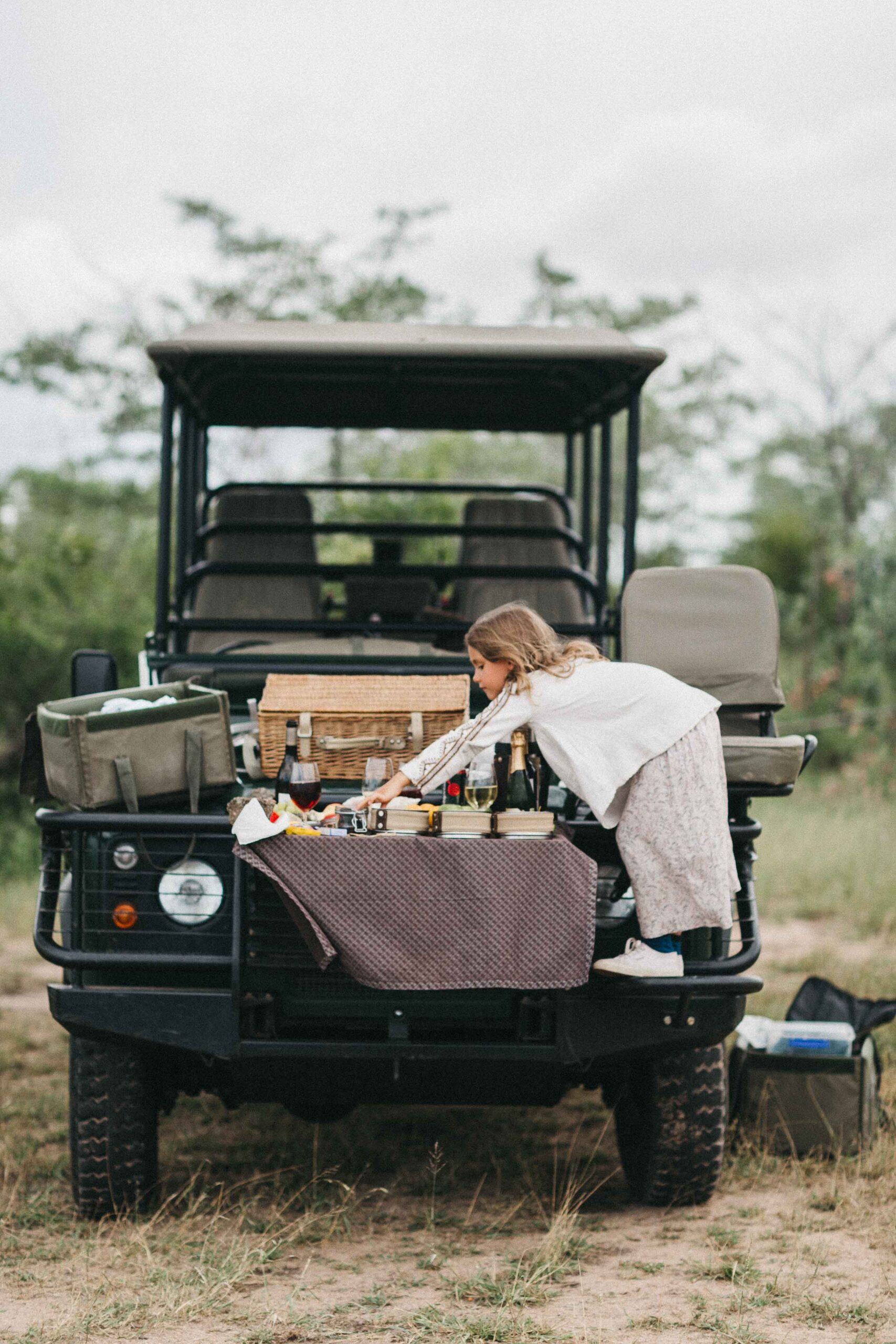 Ein junges Mädchen greift über ein Picknick auf der Motorhaube eines Safarifahrzeugs im Freien, umgeben von Graslandschaften und Bäumen. Die Szene wurde von einem erfahrenen Kinderfotografen aufgenommen und zeigt Körbe, Essen und Getränke in einer entspannten, abenteuerlichen Atmosphäre.