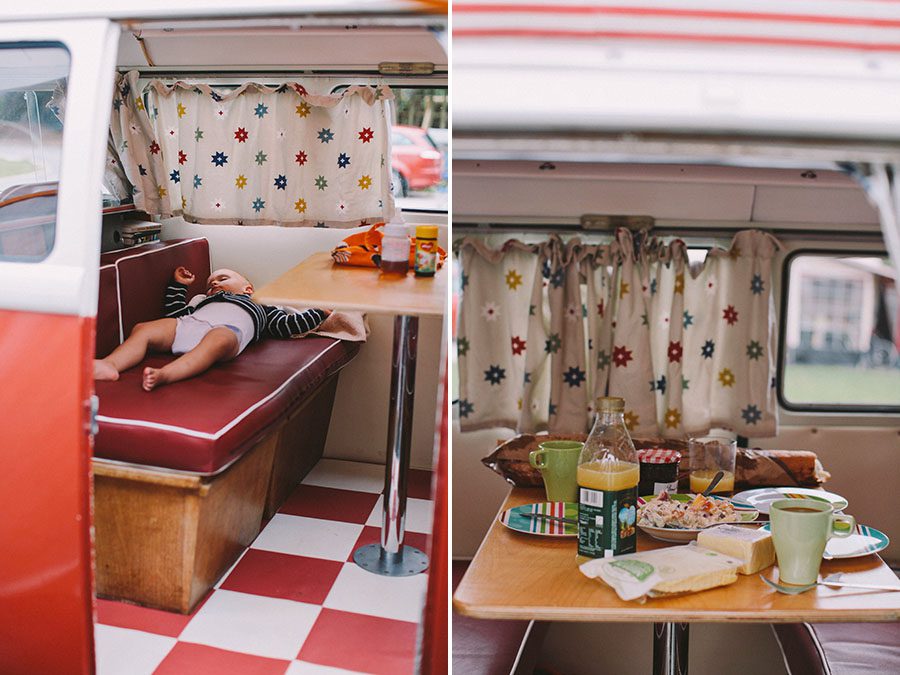 Ein Kind macht ein Nickerchen auf einem roten Sitz in einem Retro-Wohnmobil mit sterngemusterten Vorhängen. Die gemütliche Szene wurde von einem talentierten Kinderfotografen aufgenommen und zeigt Frühstücksgerichte und Getränke auf einem Tisch auf einem rot-weiß-karierten Boden.